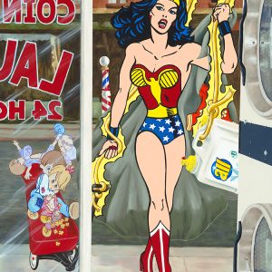 Wonder Woman carries a tarp of laundry into a laundromat, dragging detergent and a wagon full of children, set against an American street scene.